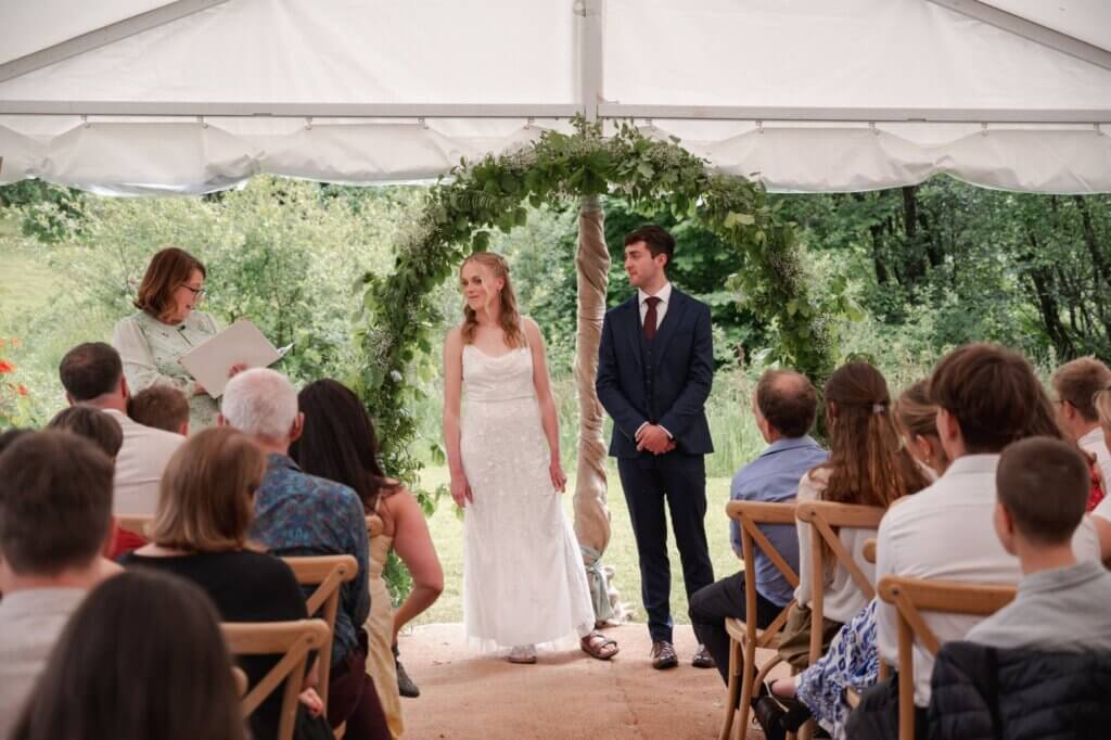 Celebrant reading from her ceremony folder as the bride and groom stand under a foliage arch in front of their guests