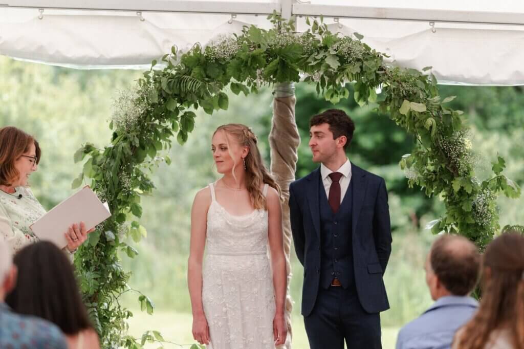 Bride and groom standing under a greenery and gypsophila arch as the celebrant reads at their garden wedding