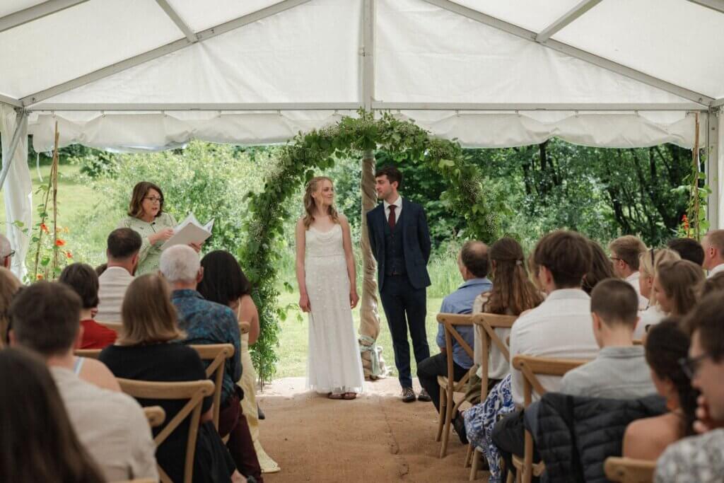 Guests seated on wooden chairs in an open-sided marquee as a couple stand under a greenery arch with the garden behind them