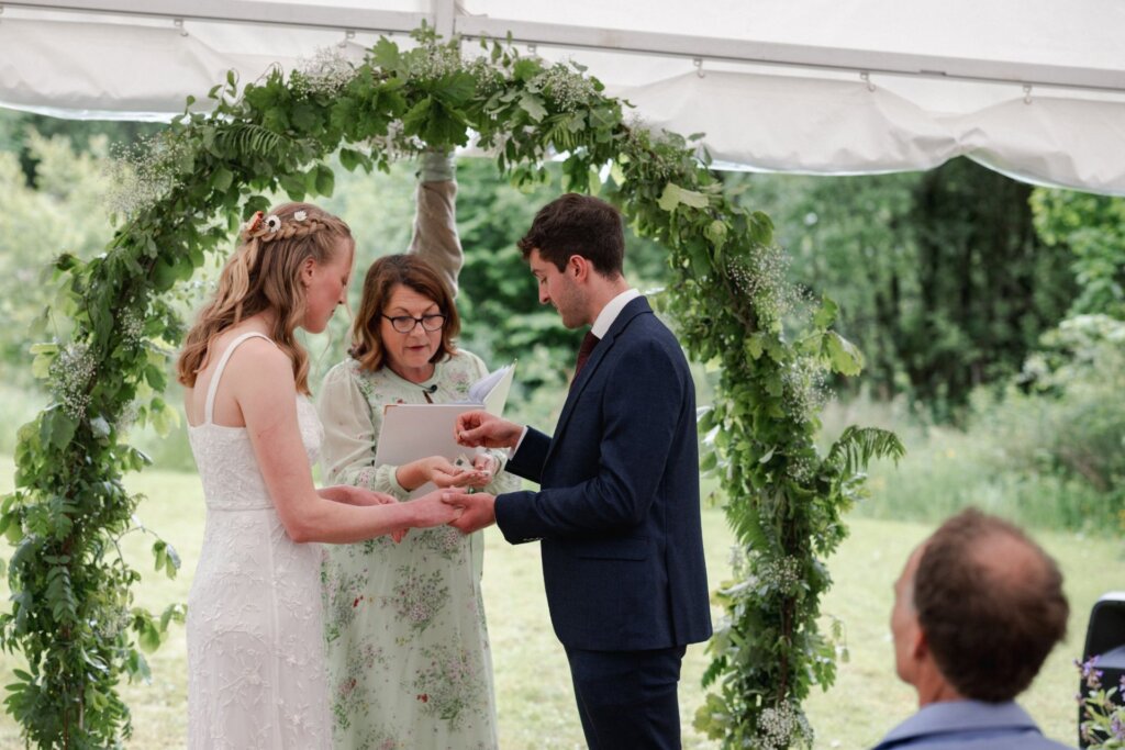 Bride and groom exchanging rings beside celebrant Jane Jones under a greenery arch in a garden marquee