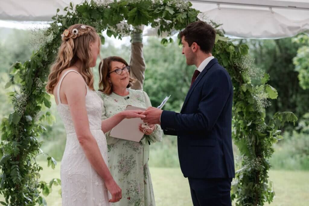 Celebrant Jane Jones smiling at a bride and groom as they exchange rings under a greenery arch