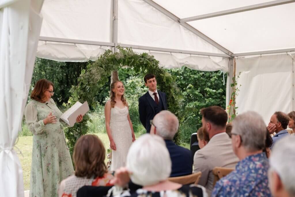View from among the guests of a celebrant-led ceremony in an open marquee with garden greenery beyond