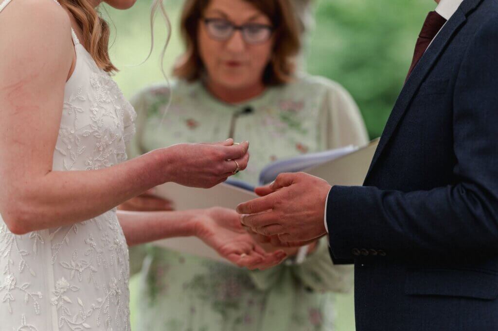 Close-up of a bride holding a wedding ring as the groom holds out his hand, with the celebrant reading behind them